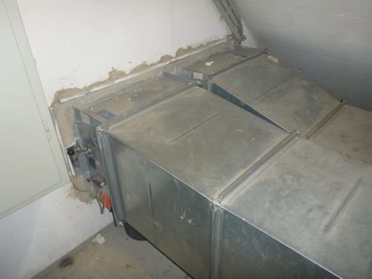 Das Bild zeigt eine große metallische Lüftungsanlage, die in einem Technikraum installiert ist. Die Anlage besteht aus mehreren rechteckigen Kanälen mit glatten Oberflächen, die an einer Wand befestigt sind. An der linken Seite sind Kabel und Anschlüsse sichtbar, während die Wand rund um die Öffnung Spuren von Abdichtmaterial aufweist.