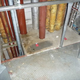 Das Bild zeigt einen Technikbereich mit mehreren senkrecht verlaufenden Rohren, die teilweise mit gelblicher Isolierung ummantelt sind. Einige Rohre sind rot lackiert, andere silbern, und sie führen durch Öffnungen im Boden, der aus Betonplatten besteht. Im Vordergrund ist ein Metallgitterboden mit Geländer zu sehen, und rechts befinden sich weitere metallische Leitungen und Bauteile, die Teil einer Lüftungs- oder Heizungsanlage sein könnten.