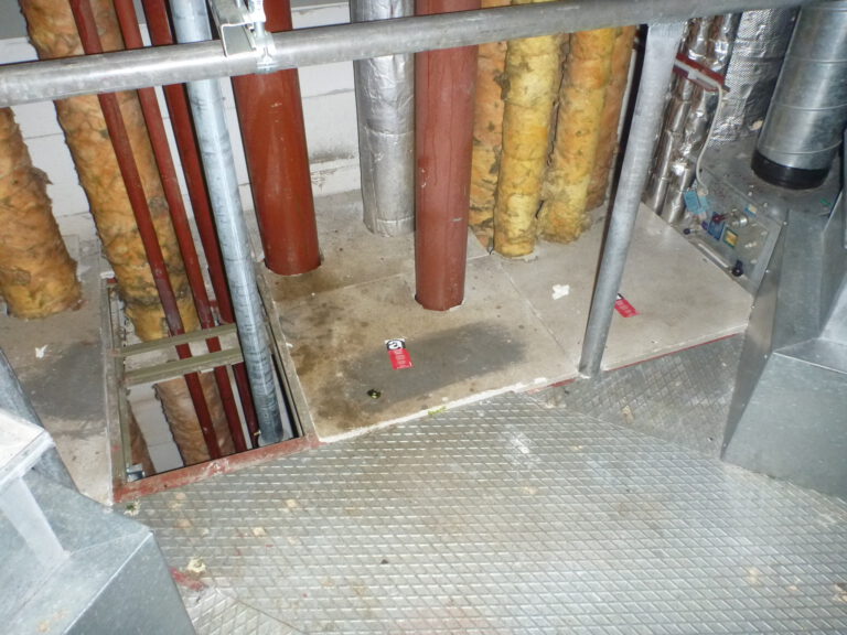 Das Bild zeigt einen Technikbereich mit mehreren senkrecht verlaufenden Rohren, die teilweise mit gelblicher Isolierung ummantelt sind. Einige Rohre sind rot lackiert, andere silbern, und sie führen durch Öffnungen im Boden, der aus Betonplatten besteht. Im Vordergrund ist ein Metallgitterboden mit Geländer zu sehen, und rechts befinden sich weitere metallische Leitungen und Bauteile, die Teil einer Lüftungs- oder Heizungsanlage sein könnten.