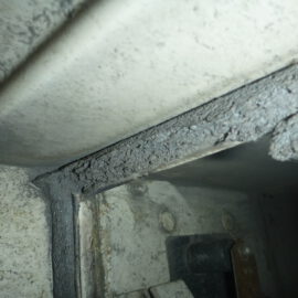 Das Bild zeigt eine Nahaufnahme des Inneren eines Lüftungskanals mit einer beschädigten Dichtung. Die Dichtung ist stark bröckelig und weist Risse sowie abgeplatzte Stellen auf, wodurch das Material unregelmäßig wirkt. Die angrenzenden Metallflächen sind verschmutzt und zeigen Staub- und Schmutzablagerungen, was auf Abnutzung und fehlende Wartung hinweist.