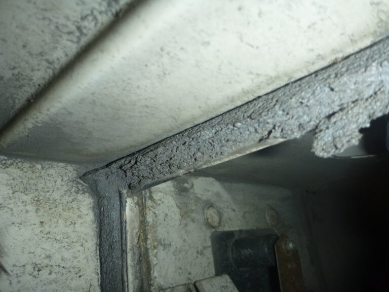 Das Bild zeigt eine Nahaufnahme des Inneren eines Lüftungskanals mit einer beschädigten Dichtung. Die Dichtung ist stark bröckelig und weist Risse sowie abgeplatzte Stellen auf, wodurch das Material unregelmäßig wirkt. Die angrenzenden Metallflächen sind verschmutzt und zeigen Staub- und Schmutzablagerungen, was auf Abnutzung und fehlende Wartung hinweist.