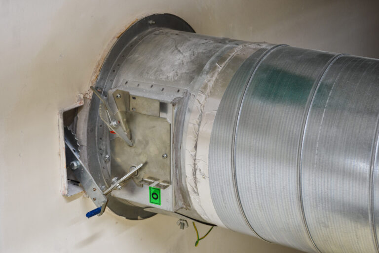 Das Bild zeigt eine metallische Lüftungsleitung, die durch eine Wand führt. An der Leitung ist eine Brandschutzklappe mit Hebelmechanismus und Befestigungselementen angebracht, die teilweise mit Dichtmaterial umgeben ist. Die Oberfläche des Kanals ist silberfarben und weist eine gerillte Struktur auf, während die Wand rund um die Öffnung leicht beschädigt wirkt.