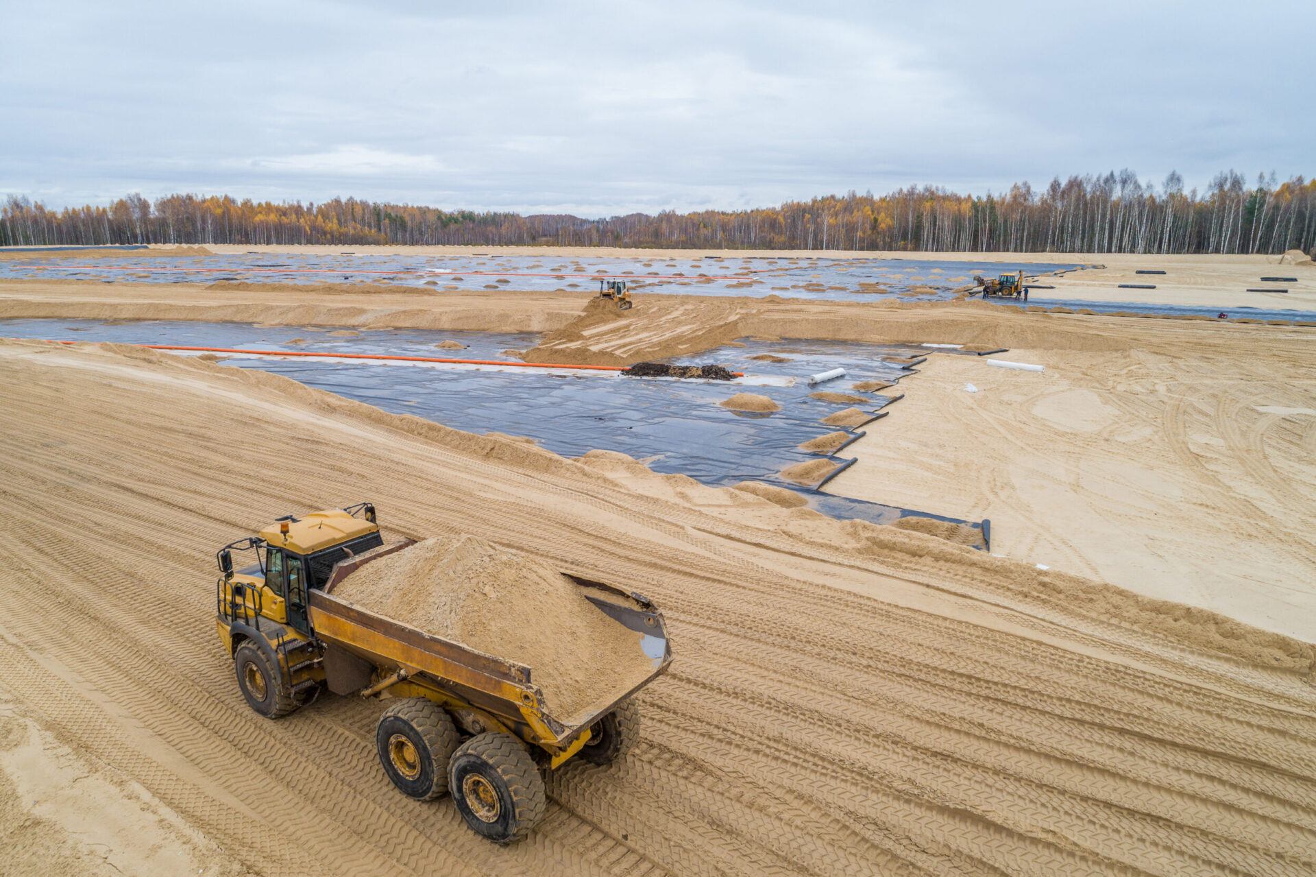 Das Bild zeigt eine große Baustelle, auf der mehrere Flächen mit schwarzer Folie abgedeckt sind, vermutlich zur Abdichtung. Im Vordergrund befindet sich ein gelbes Baustellenfahrzeug, das eine große Menge Sand transportiert, während weitere Fahrzeuge im Hintergrund arbeiten. Die Umgebung ist von hellen Sandflächen geprägt, und am Horizont erstreckt sich ein Wald mit herbstlich gefärbten Bäumen unter einem grauen, wolkenbedeckten Himmel.