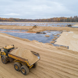 Das Bild zeigt eine große Baustelle, auf der mehrere Flächen mit schwarzer Folie abgedeckt sind, vermutlich zur Abdichtung. Im Vordergrund befindet sich ein gelbes Baustellenfahrzeug, das eine große Menge Sand transportiert, während weitere Fahrzeuge im Hintergrund arbeiten. Die Umgebung ist von hellen Sandflächen geprägt, und am Horizont erstreckt sich ein Wald mit herbstlich gefärbten Bäumen unter einem grauen, wolkenbedeckten Himmel.