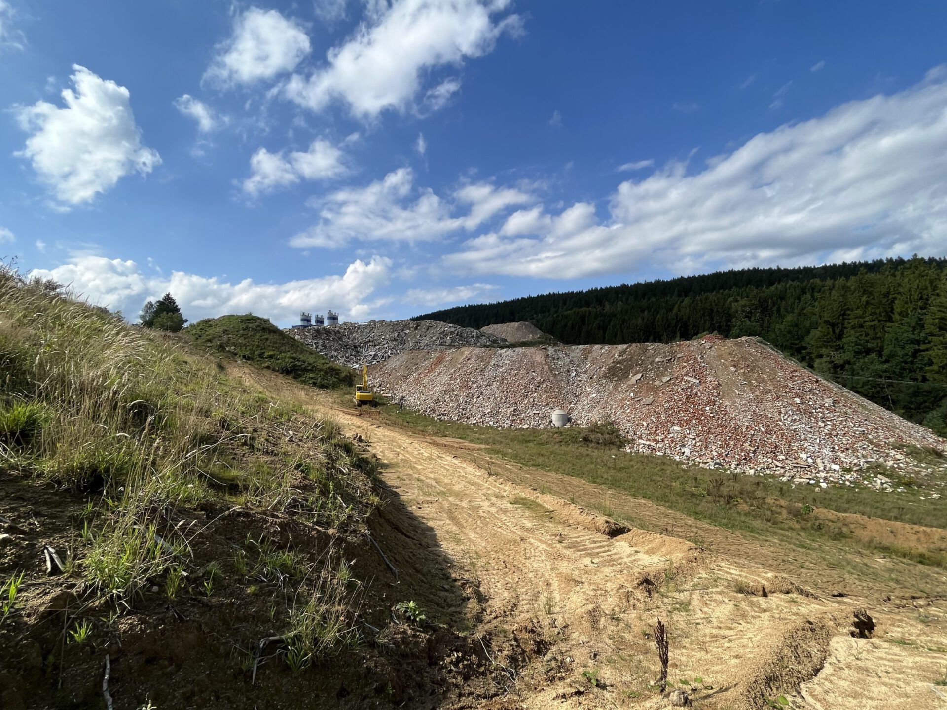 Das Bild zeigt eine offene Landschaft mit einem breiten Erdweg, der sich durch eine hügelige Fläche zieht. Links ist eine grasbewachsene Böschung zu sehen, während rechts große Haufen aus Bauschutt und Steinen liegen, die überwiegend grau und rötlich gefärbt sind. Im Hintergrund steht ein gelbes Baustellenfahrzeug nahe den Schutthaufen, dahinter erstreckt sich ein dichter Wald unter einem blauen Himmel mit vereinzelten weißen Wolken.