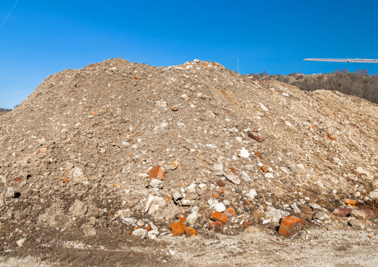 Das Bild zeigt einen großen Haufen aus Bauschutt und Erdmaterial, der sich über den gesamten Vordergrund erstreckt. Zwischen dem überwiegend grauen Material sind zahlreiche rote Ziegelstücke und helle Betonreste erkennbar. Der Hintergrund zeigt einen klaren blauen Himmel sowie eine angedeutete Landschaft mit Bäumen und einer Baukranstruktur am rechten Bildrand.