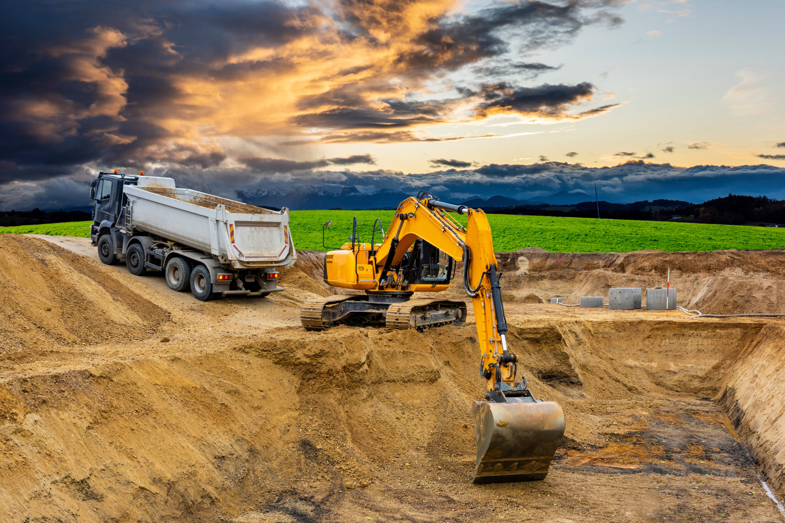 Das Bild zeigt eine Baustelle mit einem großen gelben Bagger, der in einer tiefen Erdgrube arbeitet. Daneben steht ein weißer Kipp-Lkw auf einer ebenen Fläche, bereit Material aufzunehmen oder abzuladen. Im Hintergrund erstreckt sich eine grüne Wiese, dahinter eine Bergkette unter einem dramatischen Himmel mit dunklen Wolken und orangefarbenen Lichtreflexen vom Sonnenuntergang.