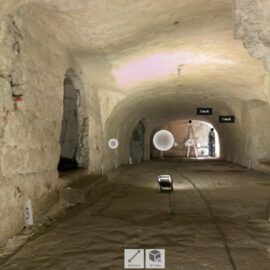 Zu sehen ist ein länglicher, tonnengewölbter Felsenkeller mit grob behauenen, hellen Gesteinswänden und -decke. Der Boden ist eben und besteht aus einer glatten, dunkleren Fläche. Entlang der Wände sind mehrere kleine Öffnungen und Nischen erkennbar. Im hinteren Bereich steht eine Person neben einem Stativ, daneben ist Baustellenbeleuchtung platziert, die den Raum ausleuchtet. Auf dem Boden verlaufen Kabel zu einem Scheinwerfer in der Bildmitte. Im Bild sind außerdem digitale Messpunkte, Markierungen („1 Kluft“, „2 Kluft“) sowie eine Mini-Kartenansicht und Bedien-Symbole eingeblendet.