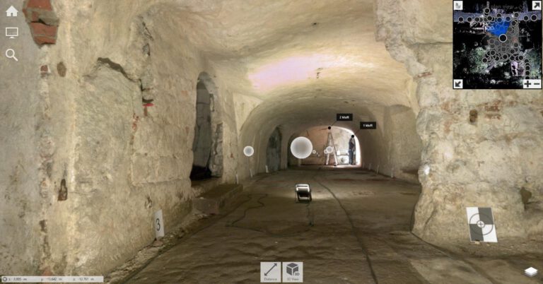 Zu sehen ist ein länglicher, tonnengewölbter Felsenkeller mit grob behauenen, hellen Gesteinswänden und -decke. Der Boden ist eben und besteht aus einer glatten, dunkleren Fläche. Entlang der Wände sind mehrere kleine Öffnungen und Nischen erkennbar. Im hinteren Bereich steht eine Person neben einem Stativ, daneben ist Baustellenbeleuchtung platziert, die den Raum ausleuchtet. Auf dem Boden verlaufen Kabel zu einem Scheinwerfer in der Bildmitte. Im Bild sind außerdem digitale Messpunkte, Markierungen („1 Kluft“, „2 Kluft“) sowie eine Mini-Kartenansicht und Bedien-Symbole eingeblendet.