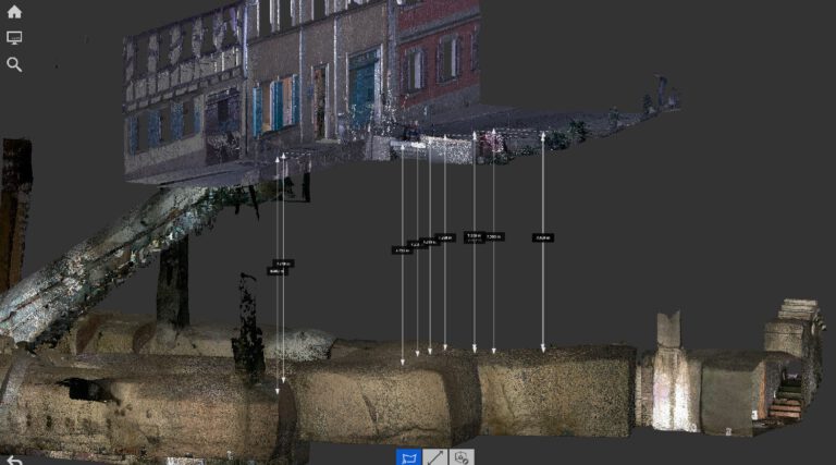 Zu sehen ist eine digitale 3D-Darstellung bzw. Punktwolkenansicht eines unterirdischen Hohlraumsystems. Oben ist ein teilweise transparent dargestellter Straßenzug mit mehreren Gebäudefassaden zu erkennen. Darunter verläuft ein länglicher, röhrenförmiger Hohlraum aus Gestein oder Beton. Mehrere senkrechte Messpfeile mit Entfernungsangaben verbinden die Oberfläche mit dem darunterliegenden Hohlraum.