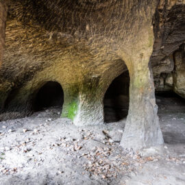 Das Foto zeigt einen weitläufigen unterirdischen Felsraum mit unregelmäßig geformten Pfeilern, die Decke und Boden miteinander verbinden. Im Hintergrund führen mehrere niedrige, dunkle Öffnungen in weitere Höhlenbereiche, während der Boden mit Erde, kleinen Steinen und vereinzelten Laubresten bedeckt ist.