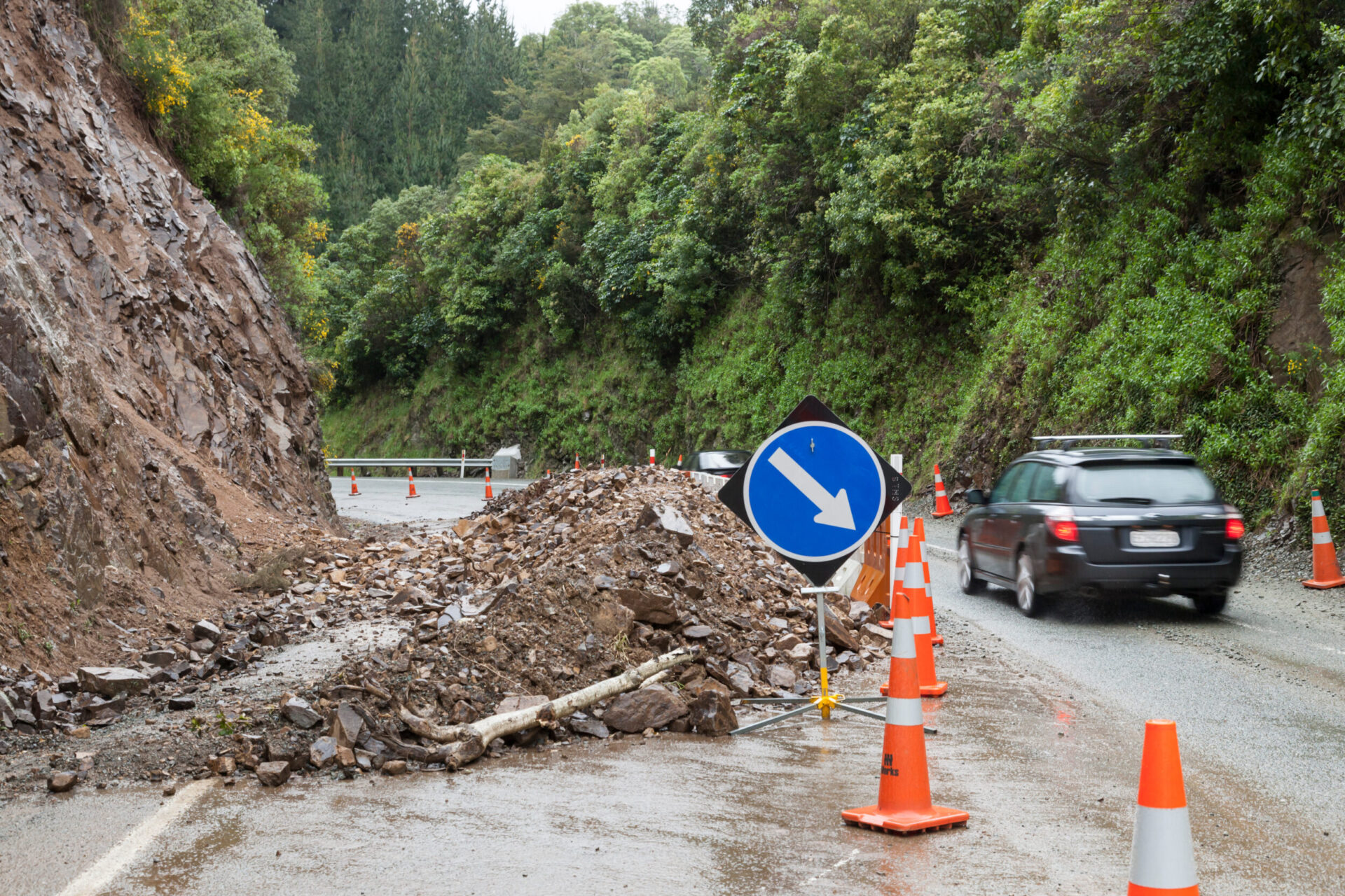 Das Bild zeigt eine Straße, die durch einen Erdrutsch teilweise blockiert ist. Auf der Fahrbahn liegt ein großer Haufen aus Steinen und Geröll, abgesichert durch mehrere orangefarbene Warnkegel und ein blaues Verkehrsschild, das die Umfahrung anzeigt. Im Hintergrund sind eine steile Felswand und dichte grüne Vegetation zu sehen, während auf der rechten Seite Fahrzeuge vorbeifahren.
