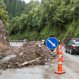 Das Bild zeigt eine Straße, die durch einen Erdrutsch teilweise blockiert ist. Auf der Fahrbahn liegt ein großer Haufen aus Steinen und Geröll, abgesichert durch mehrere orangefarbene Warnkegel und ein blaues Verkehrsschild, das die Umfahrung anzeigt. Im Hintergrund sind eine steile Felswand und dichte grüne Vegetation zu sehen, während auf der rechten Seite Fahrzeuge vorbeifahren.