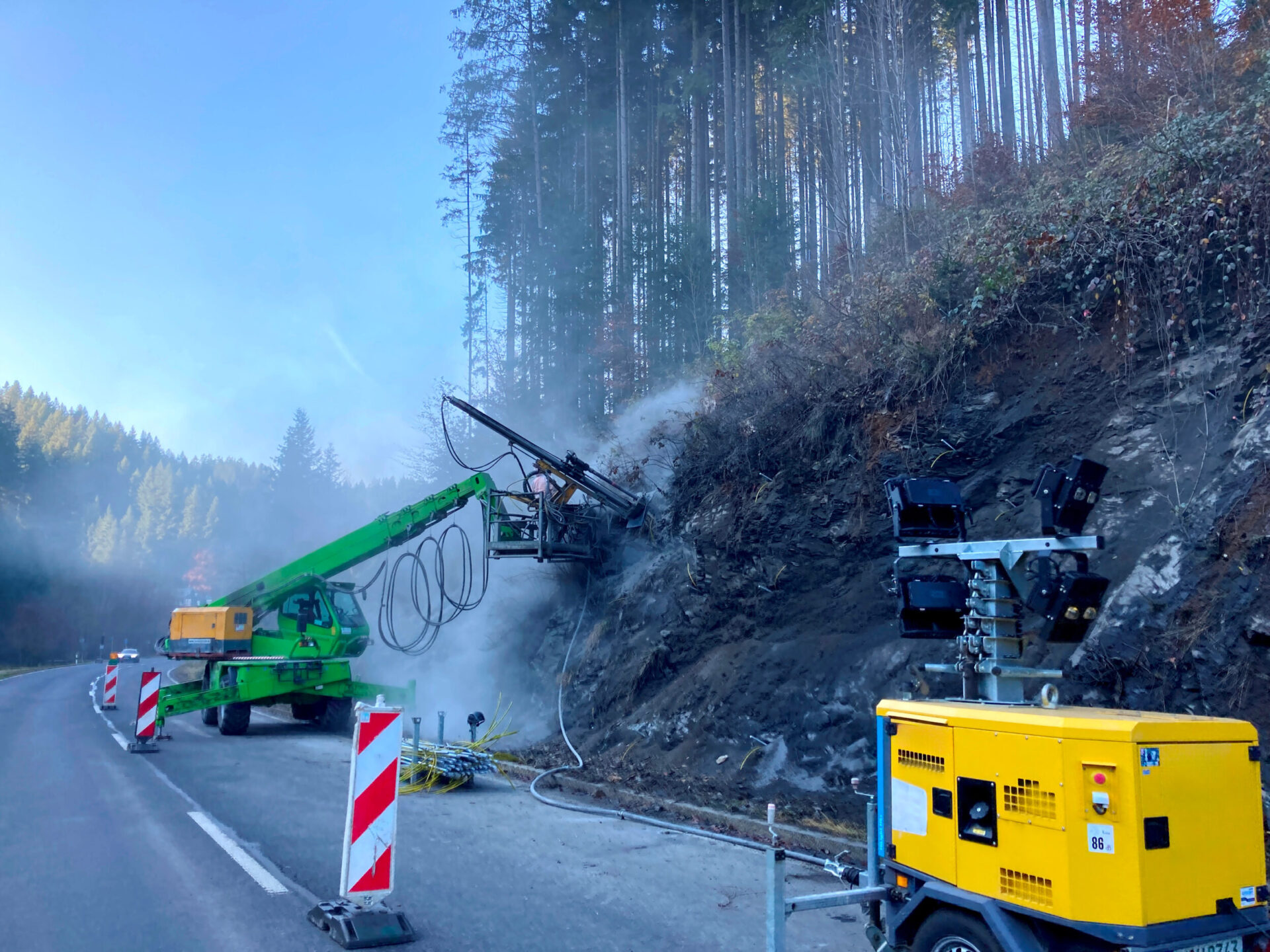 Das Bild zeigt eine Baustelle an einer kurvigen Landstraße in einem bewaldeten Tal. Rechts im Vordergrund steht ein gelber mobiler Kompressor, von dem ein Schlauch zur Bohrstelle führt. Die Straße ist durch rot-weiße Leitbaken und Warntechnik abgesichert. Links daneben befindet sich ein grünes Bohrgerät mit langem Ausleger, das direkt in die dunkle, felsige Böschung bohrt. Aus der Bohrstelle steigt Staub oder Dampf auf. Vor der Maschine steht ein gelbes Warn- und Steuergerät mit Blinklicht.