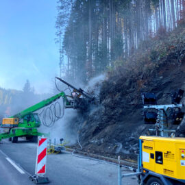 Das Bild zeigt eine Baustelle an einer kurvigen Landstraße in einem bewaldeten Tal. Rechts im Vordergrund steht ein gelber mobiler Kompressor, von dem ein Schlauch zur Bohrstelle führt. Die Straße ist durch rot-weiße Leitbaken und Warntechnik abgesichert. Links daneben befindet sich ein grünes Bohrgerät mit langem Ausleger, das direkt in die dunkle, felsige Böschung bohrt. Aus der Bohrstelle steigt Staub oder Dampf auf. Vor der Maschine steht ein gelbes Warn- und Steuergerät mit Blinklicht.