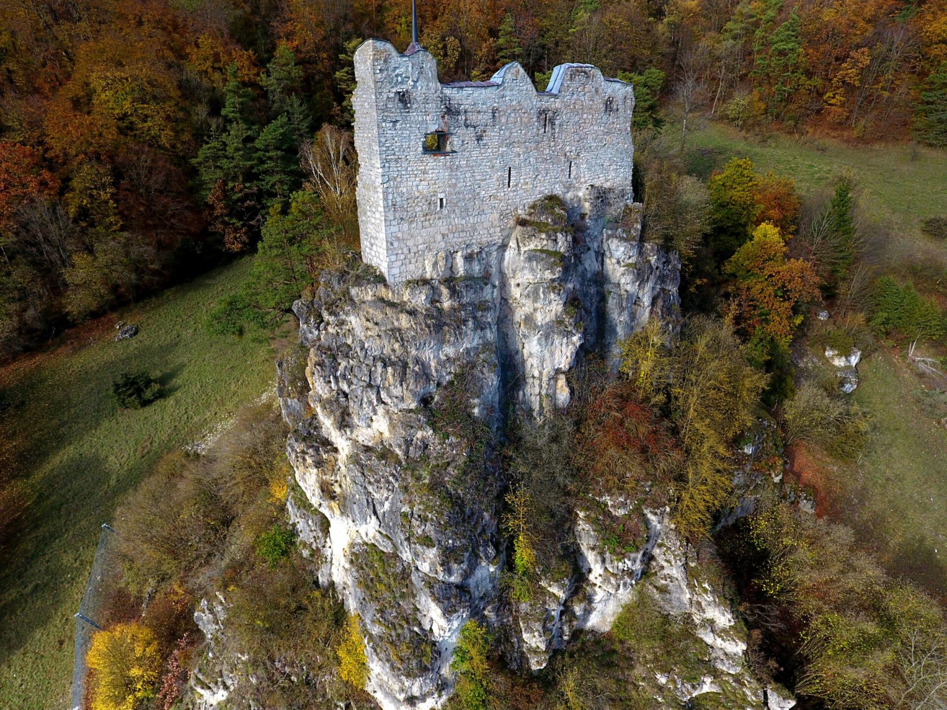 Das Bild zeigt eine alte Burgruine, die auf einem hohen, steilen Felsen thront. Die Mauern der Burg bestehen aus grauem Stein und sind teilweise beschädigt, was den historischen Charakter unterstreicht. Umgeben ist die Felsformation von herbstlichem Wald mit bunten Laubbäumen und einer grünen Wiese am Fuß des Hangs.
