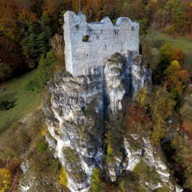 Das Bild zeigt eine alte Burgruine, die auf einem hohen, steilen Felsen thront. Die Mauern der Burg bestehen aus grauem Stein und sind teilweise beschädigt, was den historischen Charakter unterstreicht. Umgeben ist die Felsformation von herbstlichem Wald mit bunten Laubbäumen und einer grünen Wiese am Fuß des Hangs.