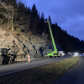 Das Bild zeigt eine nächtliche Baustellenszene an einer Landstraße entlang eines steilen bewaldeten Hangs. Rechts verläuft die zweispurige Straße, auf der mehrere Fahrzeuge mit eingeschaltetem Licht stehen oder fahren. Links die Gegenfahrbahn mit Baustellenabsicherung durch rot-weiße Leitbaken. Im Zentrum steht ein großer grüner Teleskopkran, dessen Ausleger schräg nach oben zum Hang reicht. Am Kran hängt eine Netz- bzw. Mattenkonstruktion, die am Fels befestigt wird. Auf dem Hang sind mehrere Arbeiter in orangefarbener Schutzkleidung zu erkennen, die an der Sicherung der Felswand arbeiten.
