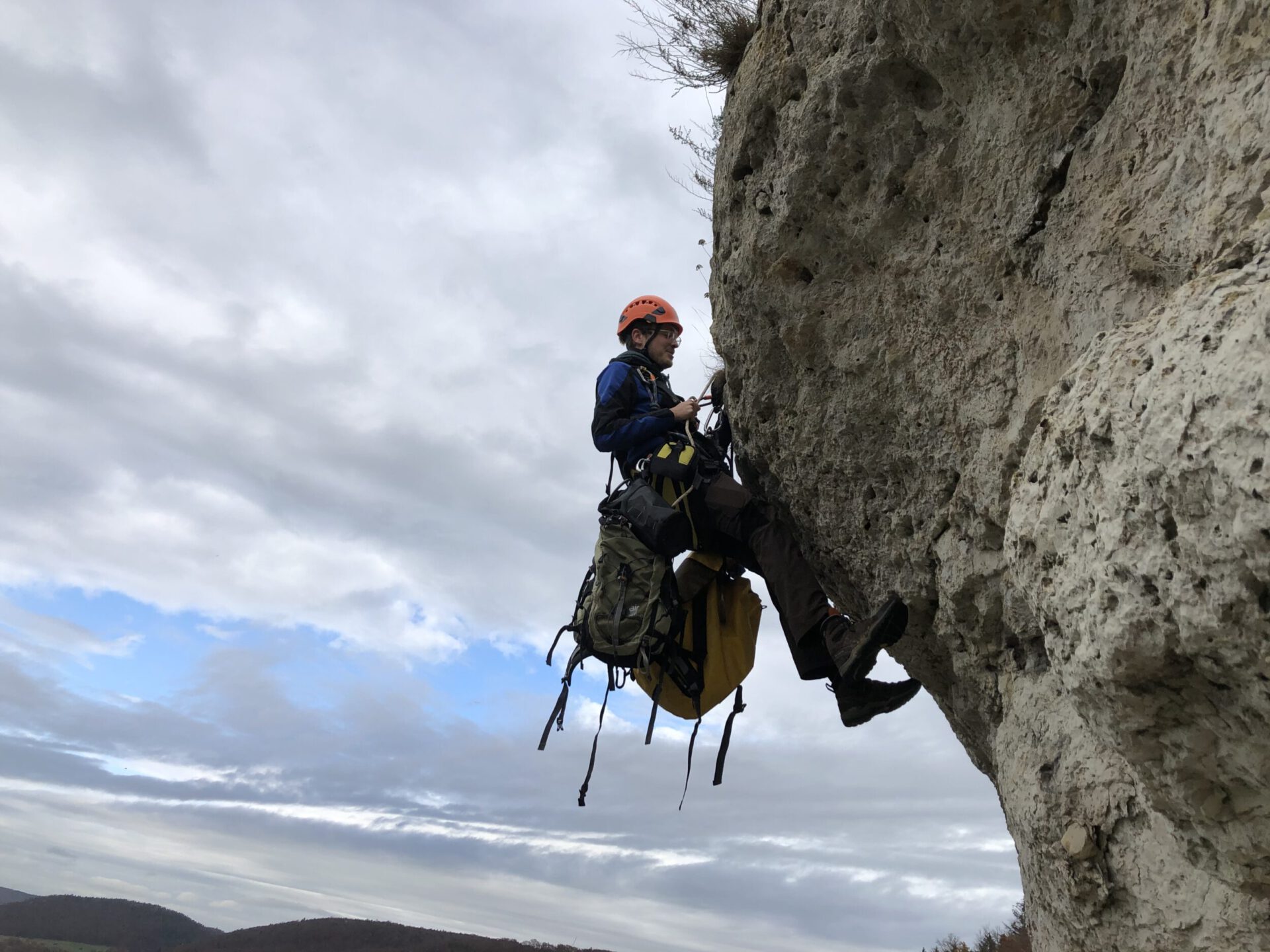 Das Bild zeigt eine Person, die mit Kletterausrüstung und Sicherheitsseilen an einer steilen Felswand hängt. Die Person trägt einen orangefarbenen Helm und hat mehrere Taschen und Ausrüstungsgegenstände am Gurt befestigt. Im Hintergrund ist ein bewölkter Himmel zu sehen, der die Szene in eine natürliche Felslandschaft einbettet.