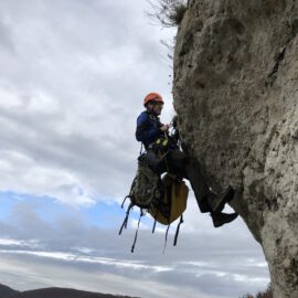 Das Bild zeigt eine Person, die mit Kletterausrüstung und Sicherheitsseilen an einer steilen Felswand hängt. Die Person trägt einen orangefarbenen Helm und hat mehrere Taschen und Ausrüstungsgegenstände am Gurt befestigt. Im Hintergrund ist ein bewölkter Himmel zu sehen, der die Szene in eine natürliche Felslandschaft einbettet.