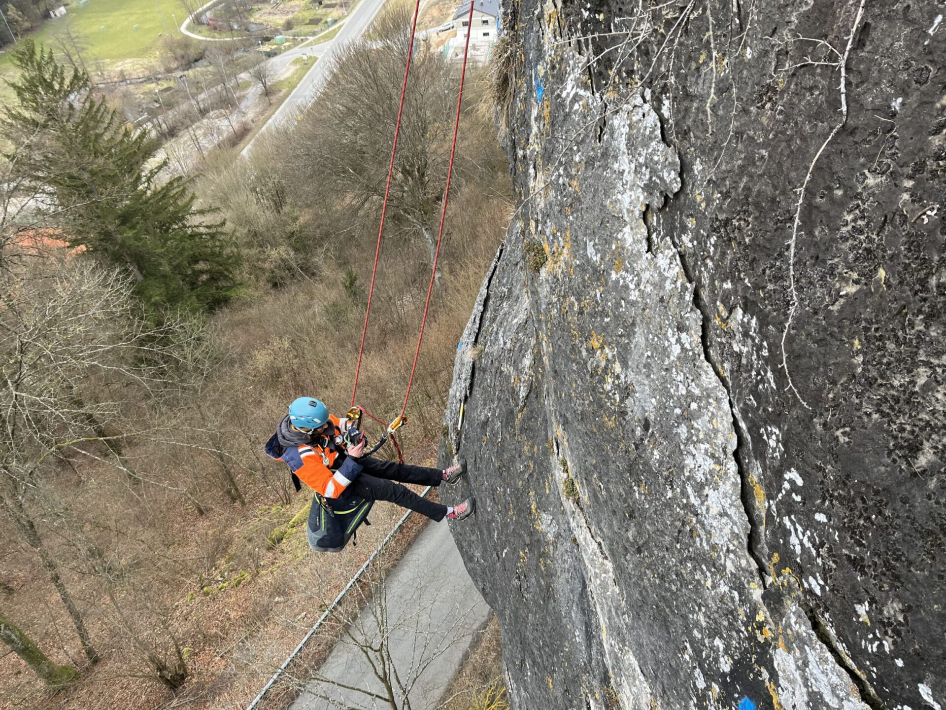 Das Bild zeigt eine Person, die mit Kletterausrüstung und Sicherheitsseilen an einer steilen Felswand hängt. Die Person trägt einen Helm und eine orangefarbene Schutzweste und arbeitet in großer Höhe über einer Straße, die im Hintergrund sichtbar ist. Umgeben ist die Szene von kahlen Bäumen und einer hügeligen Landschaft mit vereinzelten Grünflächen.