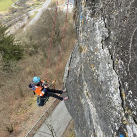 Das Bild zeigt eine Person, die mit Kletterausrüstung und Sicherheitsseilen an einer steilen Felswand hängt. Die Person trägt einen Helm und eine orangefarbene Schutzweste und arbeitet in großer Höhe über einer Straße, die im Hintergrund sichtbar ist. Umgeben ist die Szene von kahlen Bäumen und einer hügeligen Landschaft mit vereinzelten Grünflächen.