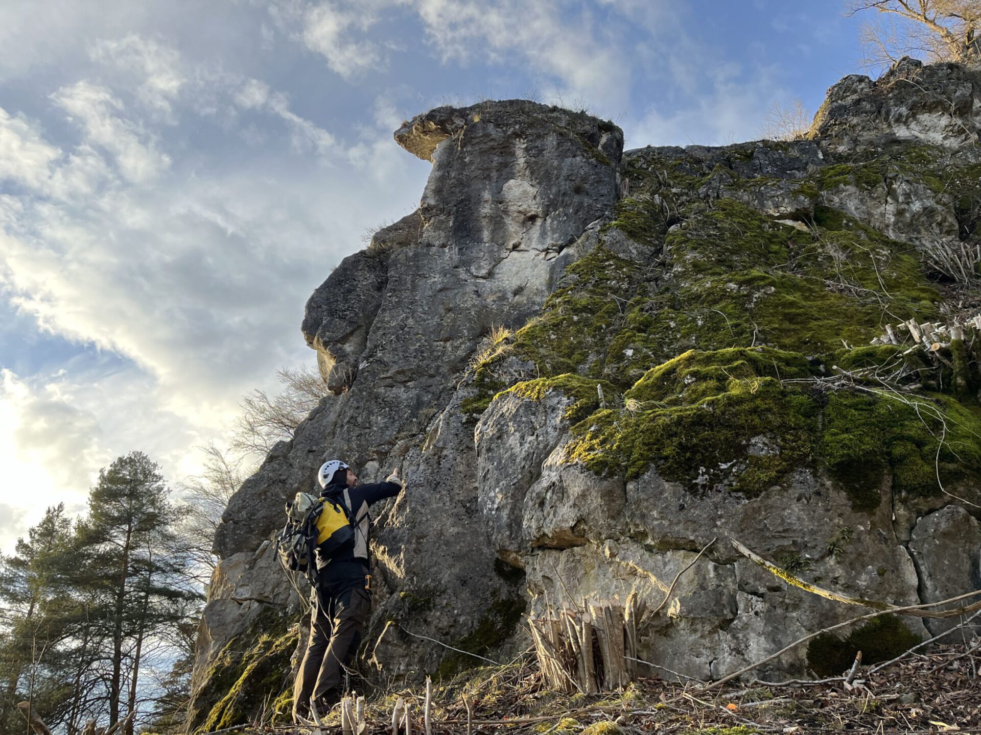 Das Bild zeigt eine große Felsformation, die steil in die Höhe ragt und teilweise mit grünem Moos bewachsen ist. Im Vordergrund steht eine Person mit Helm und Rucksack, die den Felsen betrachtet oder fotografiert. Der Himmel ist leicht bewölkt und von Sonnenlicht erhellt, wodurch die Szene eine natürliche und offene Landschaft vermittelt.