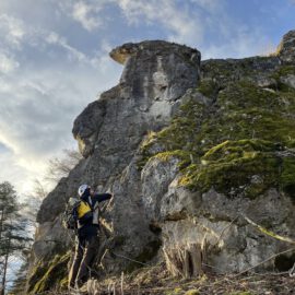 Das Bild zeigt eine große Felsformation, die steil in die Höhe ragt und teilweise mit grünem Moos bewachsen ist. Im Vordergrund steht eine Person mit Helm und Rucksack, die den Felsen betrachtet oder fotografiert. Der Himmel ist leicht bewölkt und von Sonnenlicht erhellt, wodurch die Szene eine natürliche und offene Landschaft vermittelt.