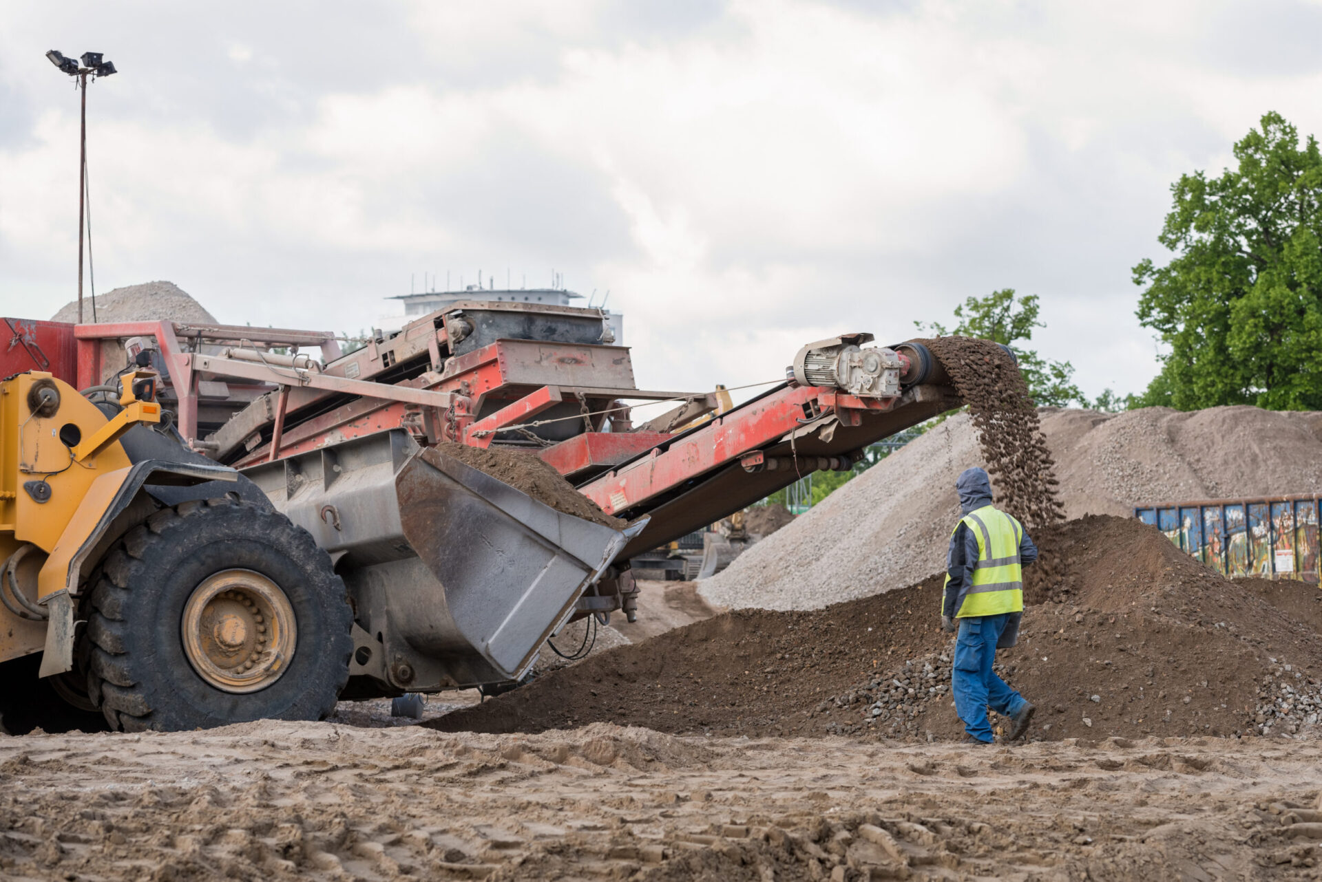 Das Bild zeigt eine Baustelle mit einer großen Maschine, die über ein Förderband Erde und Steine auf einen Haufen ablädt. Links ist ein gelbes Baustellenfahrzeug zu sehen, während im Vordergrund eine Person mit Warnweste und Mütze neben dem Materialhaufen steht. Im Hintergrund befinden sich weitere Erdhügel und grüne Bäume unter einem bewölkten Himmel.
