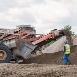 Das Bild zeigt eine Baustelle mit einer großen Maschine, die über ein Förderband Erde und Steine auf einen Haufen ablädt. Links ist ein gelbes Baustellenfahrzeug zu sehen, während im Vordergrund eine Person mit Warnweste und Mütze neben dem Materialhaufen steht. Im Hintergrund befinden sich weitere Erdhügel und grüne Bäume unter einem bewölkten Himmel.