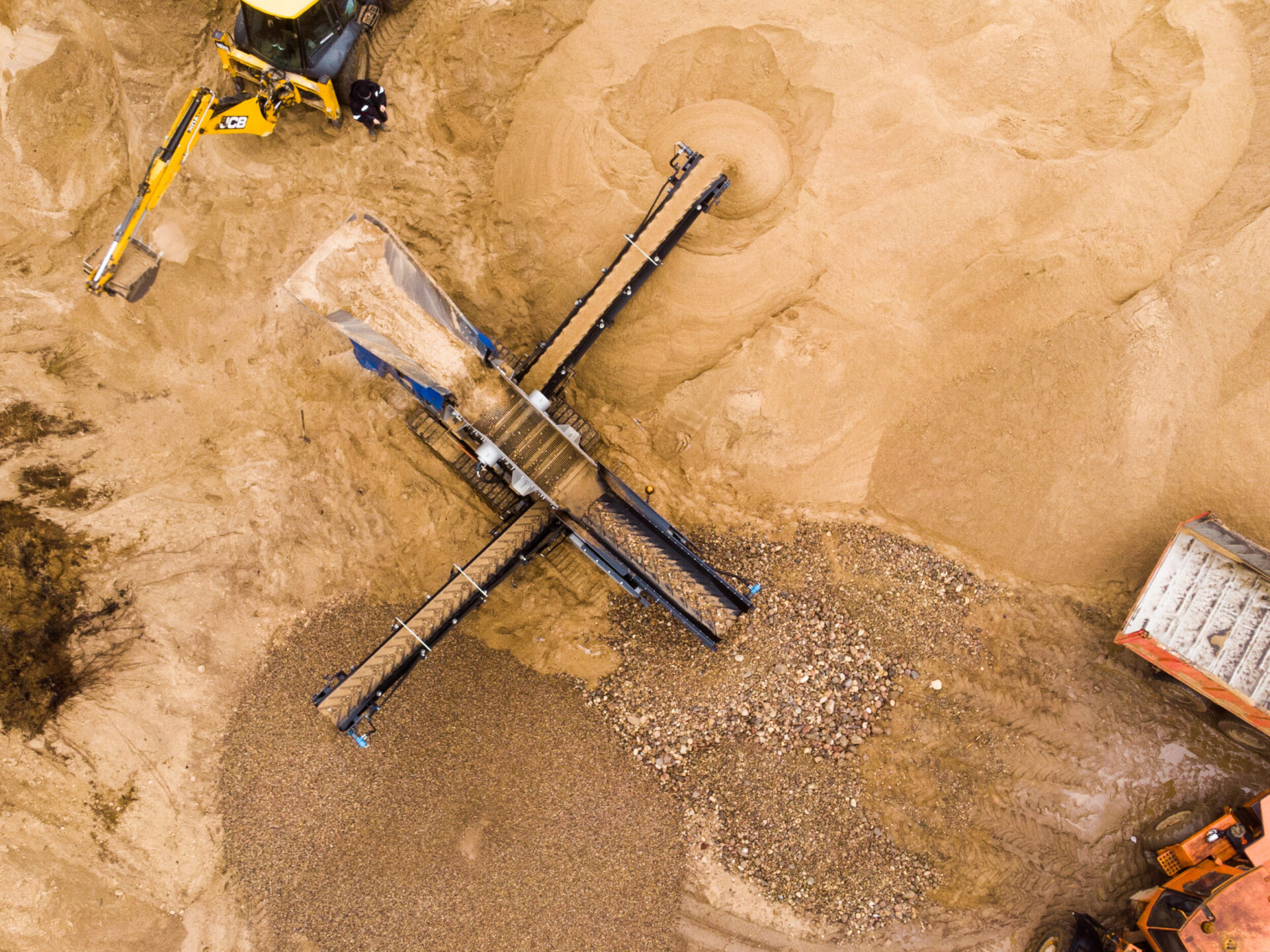 Das Bild zeigt eine Baustelle mit einer großen Maschine zur Materialaufbereitung, die von oben fotografiert wurde. Die Maschine verfügt über mehrere Förderbänder, die Sand und Kies sortieren, wobei ein großer Haufen aus grobem Material im unteren Bereich sichtbar ist. Rund um die Maschine liegen helle Sandflächen mit kreisförmigen Spuren, und am oberen sowie rechten Bildrand sind Teile von Baustellenfahrzeugen zu erkennen.