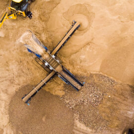 Das Bild zeigt eine Baustelle mit einer großen Maschine zur Materialaufbereitung, die von oben fotografiert wurde. Die Maschine verfügt über mehrere Förderbänder, die Sand und Kies sortieren, wobei ein großer Haufen aus grobem Material im unteren Bereich sichtbar ist. Rund um die Maschine liegen helle Sandflächen mit kreisförmigen Spuren, und am oberen sowie rechten Bildrand sind Teile von Baustellenfahrzeugen zu erkennen.