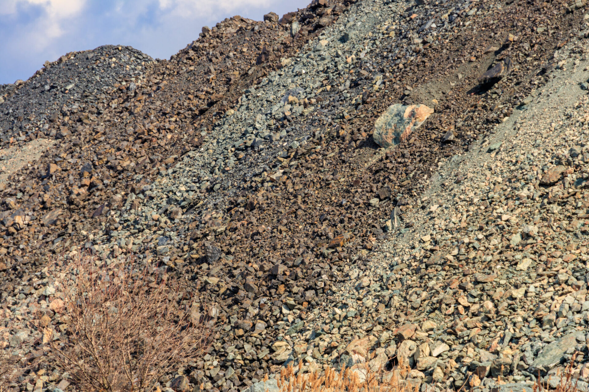 Das Bild zeigt einen großen Haufen aus lockerem Gestein und Geröll, der sich über den gesamten Bildbereich erstreckt. Die Steine variieren in Größe und Farbe, von kleinen dunklen Brocken bis zu größeren, helleren Felsen, die vereinzelt hervortreten. Im Vordergrund sind einige trockene Pflanzen zu sehen, während der Hintergrund von weiteren Geröllhängen und einem leicht bewölkten Himmel geprägt ist.