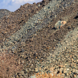 Das Bild zeigt einen großen Haufen aus lockerem Gestein und Geröll, der sich über den gesamten Bildbereich erstreckt. Die Steine variieren in Größe und Farbe, von kleinen dunklen Brocken bis zu größeren, helleren Felsen, die vereinzelt hervortreten. Im Vordergrund sind einige trockene Pflanzen zu sehen, während der Hintergrund von weiteren Geröllhängen und einem leicht bewölkten Himmel geprägt ist.