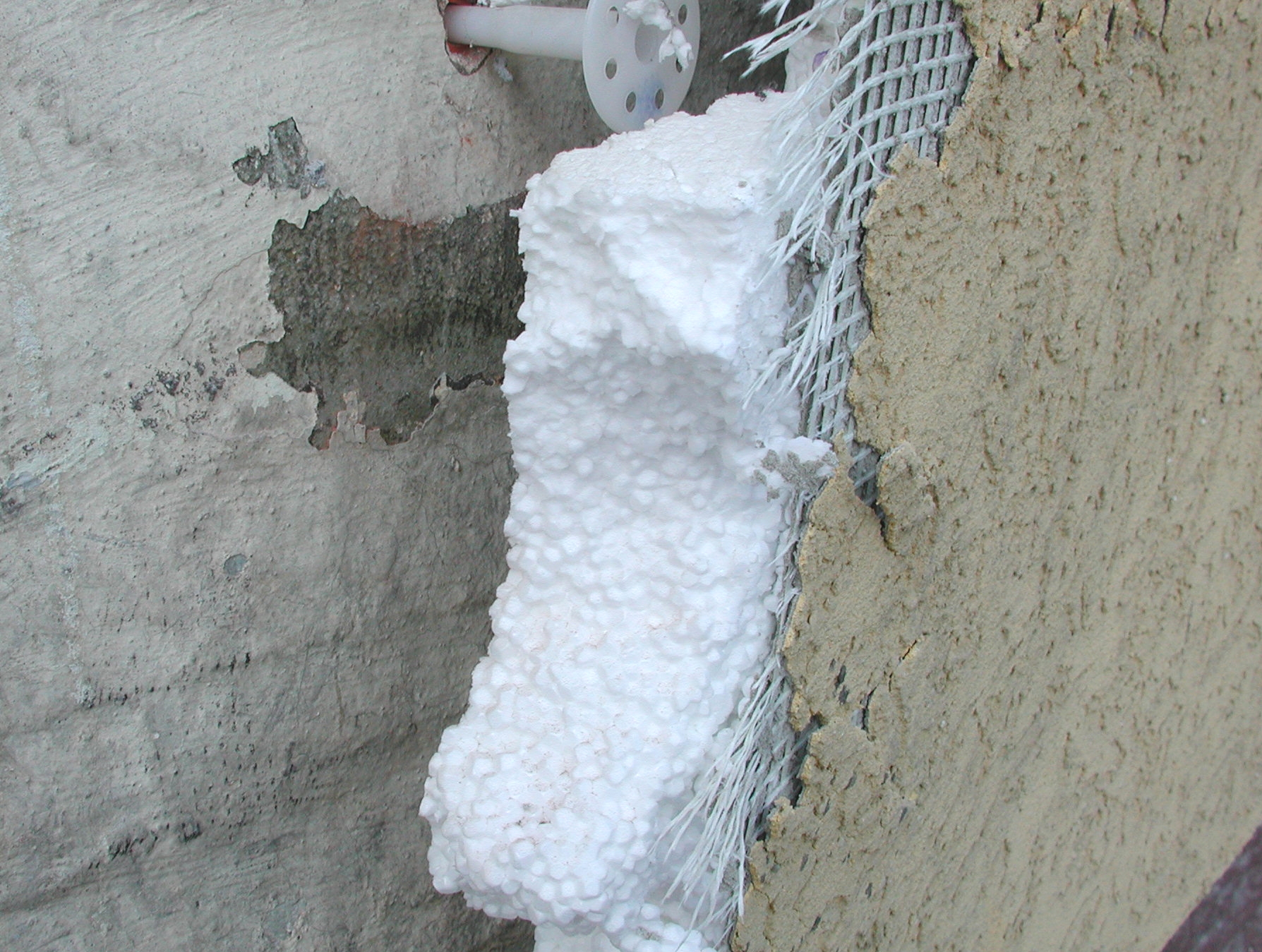 Zu sehen ist ein aufgeschnittener oder geöffneter Wandaufbau an einer Gebäudeecke. In der Mitte befindet sich ein weißes, körnig strukturiertes Dämmmaterial, das senkrecht zwischen zwei Wandflächen liegt. Rechts davon ist ein Armierungsgewebe aus feinem, weißem Kunststoffgeflecht sichtbar, das teilweise von Putz umgeben ist.