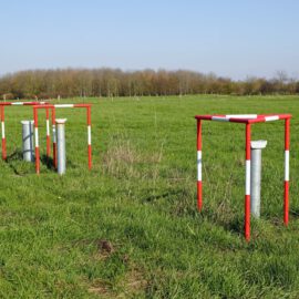 Das Bild zeigt eine grüne Wiese mit mehreren silberfarbenen Rohren, die senkrecht aus dem Boden ragen. Um jedes Rohr ist eine rot-weiß markierte Schutzkonstruktion aus Metall angebracht, die wie ein rechteckiger Rahmen aussieht. Im Hintergrund sind Bäume und ein klarer blauer Himmel zu sehen, was auf eine offene Landschaft hinweist.