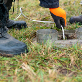 Das Bild zeigt eine Nahaufnahme von einer Messung auf einer Wiese. Im Vordergrund sind zwei Metallringe in einer rechteckigen Halterung zu sehen, die in den Boden eingelassen sind. Eine Person trägt orangefarbene Arbeitshandschuhe und hält ein Messband in einen der Ringe, während daneben robuste Arbeitsstiefel auf dem Gras stehen.