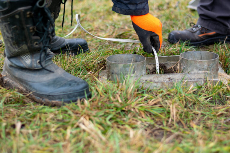 Das Bild zeigt eine Nahaufnahme von einer Messung auf einer Wiese. Im Vordergrund sind zwei Metallringe in einer rechteckigen Halterung zu sehen, die in den Boden eingelassen sind. Eine Person trägt orangefarbene Arbeitshandschuhe und hält ein Messband in einen der Ringe, während daneben robuste Arbeitsstiefel auf dem Gras stehen.