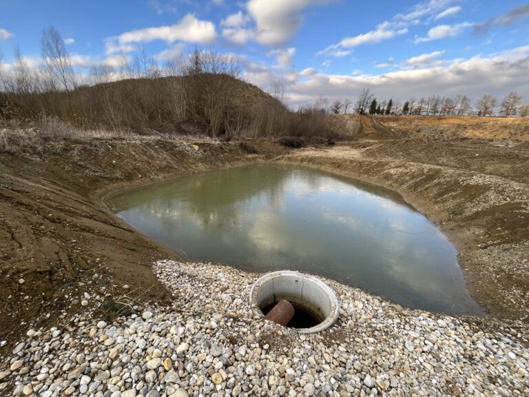 Das Bild zeigt ein künstlich angelegtes Wasserbecken in einer offenen Erdlandschaft. Im Vordergrund befindet sich ein runder Betonrohranschluss mit einem Metallrohr darin, umgeben von Kieselsteinen. Das Wasser im Becken ist grünlich und wird von unbewachsenen Erdhängen eingefasst, während im Hintergrund Bäume und ein leicht bewölkter Himmel zu sehen sind.
