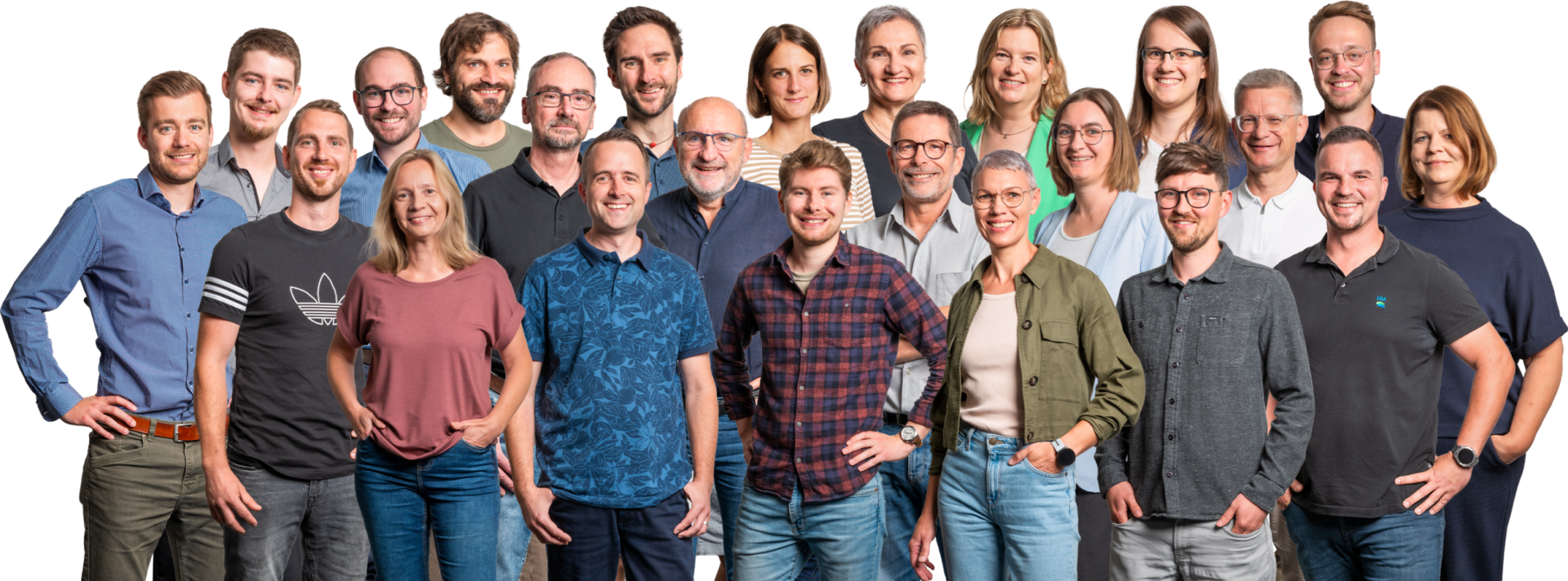 Das Bild zeigt eine größere Gruppe von Personen, die für ein Gruppenfoto posiert. Die Personen stehen in mehreren Reihen vor einem weißen Hintergrund und tragen unterschiedliche Freizeit- und Business-Kleidung wie Hemden, T-Shirts und Blusen in verschiedenen Farben und Mustern. Einige Personen haben die Hände in den Taschen oder an der Hüfte, was dem Bild eine lockere und ungezwungene Atmosphäre verleiht.