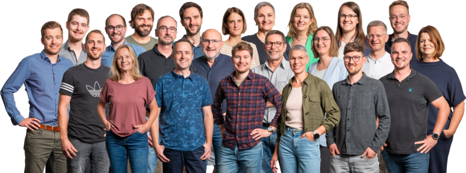 Das Bild zeigt eine größere Gruppe von Personen, die für ein Gruppenfoto posiert. Die Personen stehen in mehreren Reihen vor einem weißen Hintergrund und tragen unterschiedliche Freizeit- und Business-Kleidung wie Hemden, T-Shirts und Blusen in verschiedenen Farben und Mustern. Einige Personen haben die Hände in den Taschen oder an der Hüfte, was dem Bild eine lockere und ungezwungene Atmosphäre verleiht.
