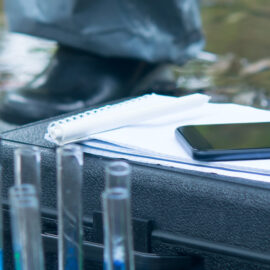 Das Bild zeigt eine Nahaufnahme von einem mobilen Labor-Setup im Freien, vermutlich an einem Gewässerrand. Im Vordergrund stehen mehrere Glasflaschen und Reagenzgläser, teilweise mit farbigen Flüssigkeiten gefüllt, sowie eine schwarze Transportbox mit darauf liegenden Notizblättern und einem Smartphone. Eine Hand mit blauer Schutzhandschuhen berührt die Unterlagen, während im Hintergrund unscharf Wasser und ein Schuh auf steinigem Boden zu erkennen sind.