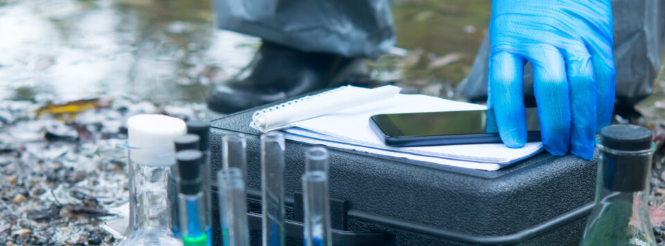 Das Bild zeigt eine Nahaufnahme von einem mobilen Labor-Setup im Freien, vermutlich an einem Gewässerrand. Im Vordergrund stehen mehrere Glasflaschen und Reagenzgläser, teilweise mit farbigen Flüssigkeiten gefüllt, sowie eine schwarze Transportbox mit darauf liegenden Notizblättern und einem Smartphone. Eine Hand mit blauer Schutzhandschuhen berührt die Unterlagen, während im Hintergrund unscharf Wasser und ein Schuh auf steinigem Boden zu erkennen sind.