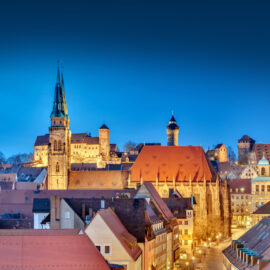 Das Bild zeigt eine Panoramaansicht der Nürnberger Altstadt bei Abenddämmerung. Im Vordergrund sind die Dächer historischer Gebäude zu sehen, während im Hintergrund die Kaiserburg mit ihren markanten Türmen und Mauern hervorsticht.