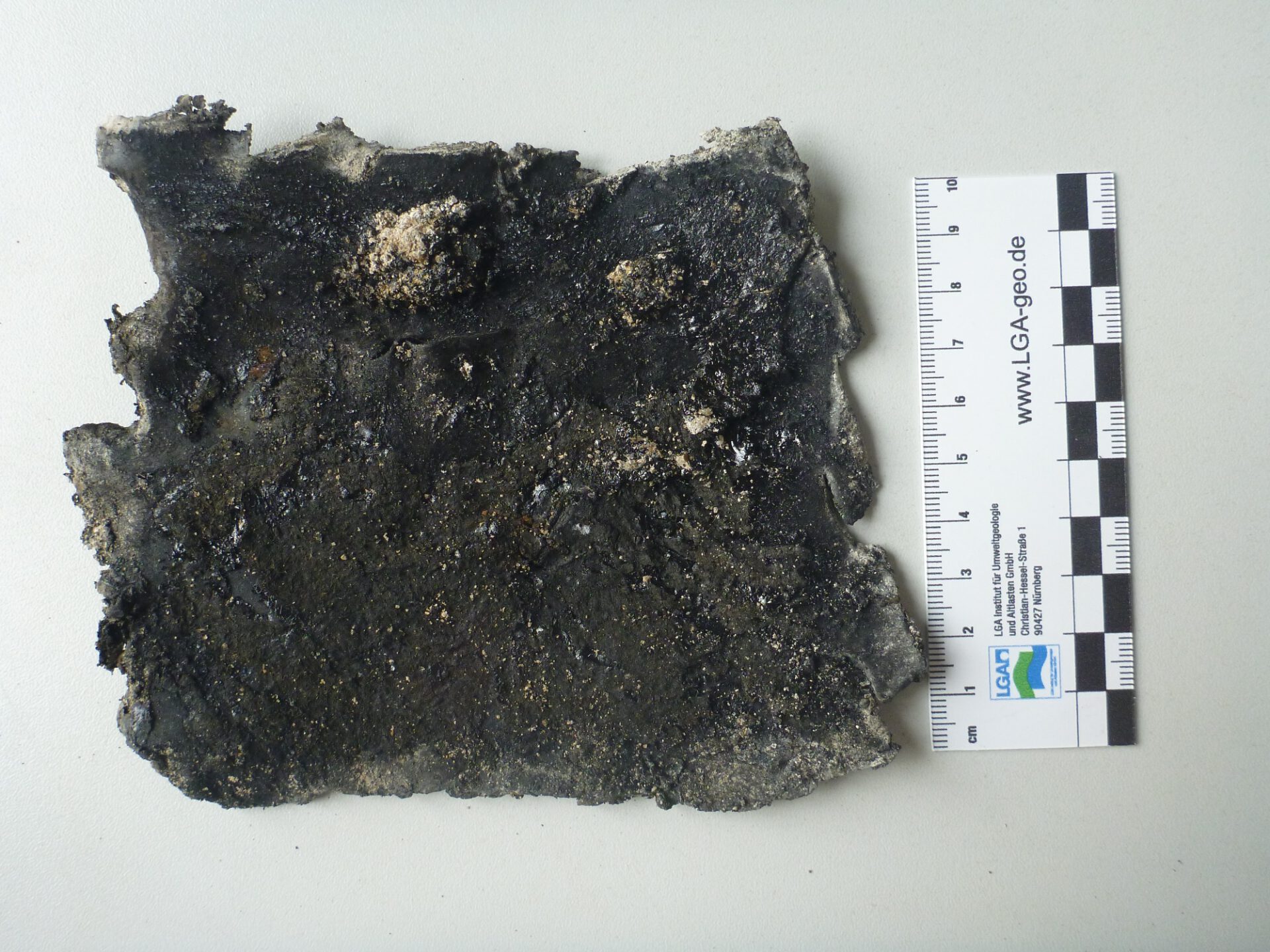 Zu sehen ist ein flaches, unregelmäßig geformtes, schwarz-dunkelgraues Materialstück auf hellem, glattem Untergrund. Die Oberfläche wirkt rau, teilweise glänzend und körnig, mit eingelagerten helleren, sandigen Partikeln.