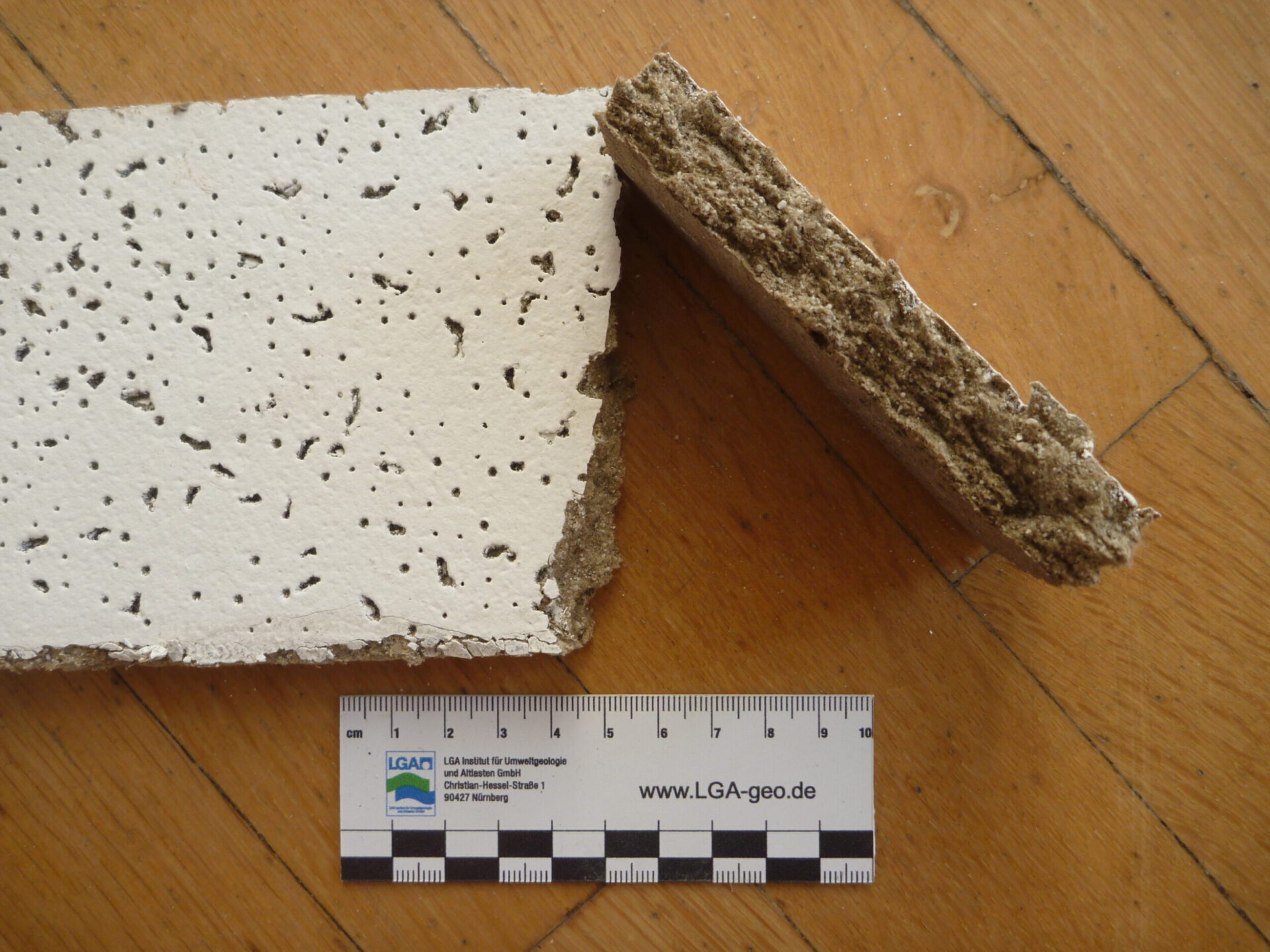 Auf dem Bild sind zwei Bruchstücke eines Baumaterials auf einem Holzfußboden zu sehen. Das Material wirkt insgesamt wie eine Deckenplatte mit poröser Struktur.