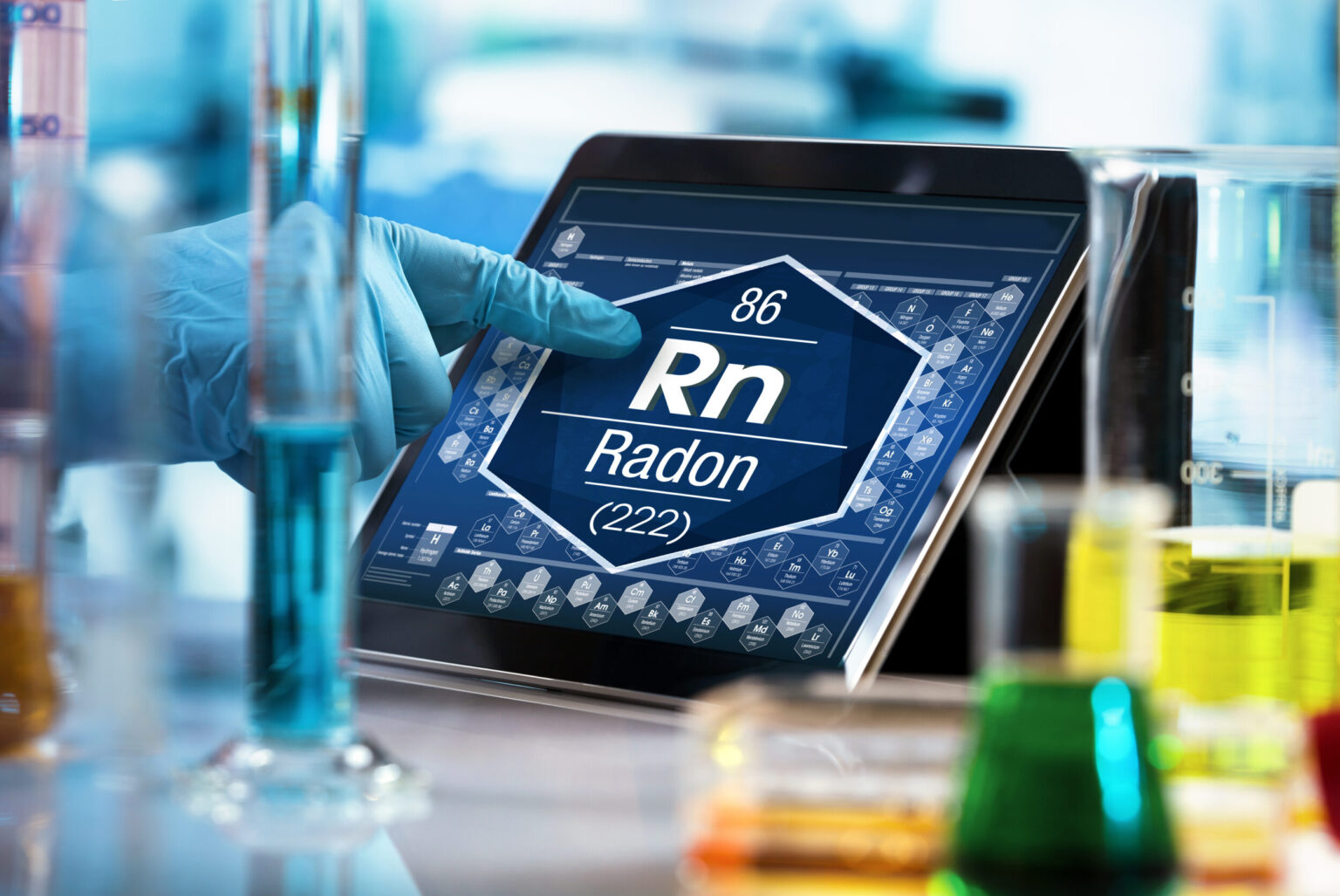 Das Bild zeigt eine Laborumgebung mit mehreren Messzylindern und Bechergläsern, die mit unterschiedlich gefärbten Flüssigkeiten gefüllt sind. Im Hintergrund wird auf einem Tablet eine digitale Darstellung eines chemischen Elements angezeigt, während eine behandschuhte Hand auf den Bildschirm zeigt.