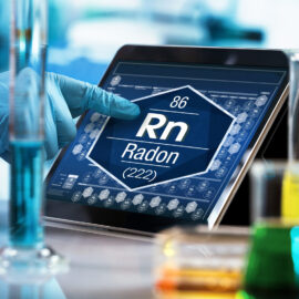 Das Bild zeigt eine Laborumgebung mit mehreren Messzylindern und Bechergläsern, die mit unterschiedlich gefärbten Flüssigkeiten gefüllt sind. Im Hintergrund wird auf einem Tablet eine digitale Darstellung eines chemischen Elements angezeigt, während eine behandschuhte Hand auf den Bildschirm zeigt.