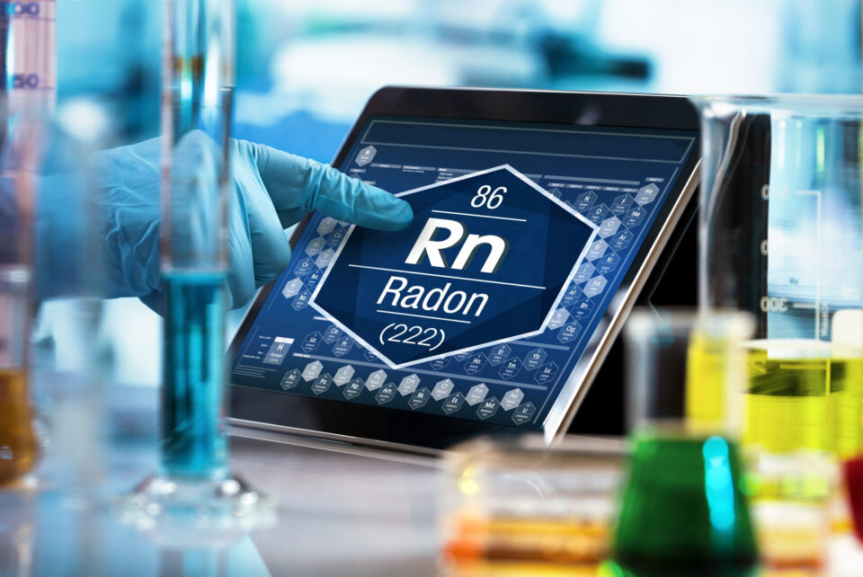 Das Bild zeigt eine Laborumgebung mit mehreren Messzylindern und Bechergläsern, die mit unterschiedlich gefärbten Flüssigkeiten gefüllt sind. Im Hintergrund wird auf einem Tablet eine digitale Darstellung eines chemischen Elements angezeigt, während eine behandschuhte Hand auf den Bildschirm zeigt.
