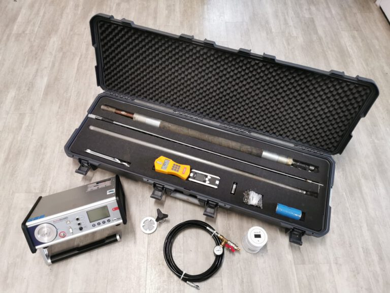 Das Bild zeigt eine geöffnete Transportbox mit verschiedenen Mess- und Bohrwerkzeugen, die ordentlich in Schaumstoffeinsätzen liegen. Darin befinden sich lange Metallstangen, ein gelbes Handgerät sowie kleinere Zubehörteile. Vor der Box sind ein silbernes Messgerät, ein schwarzes Kabel mit Anschlüssen und weitere kleine Komponenten auf einem hellen Boden ausgelegt.
